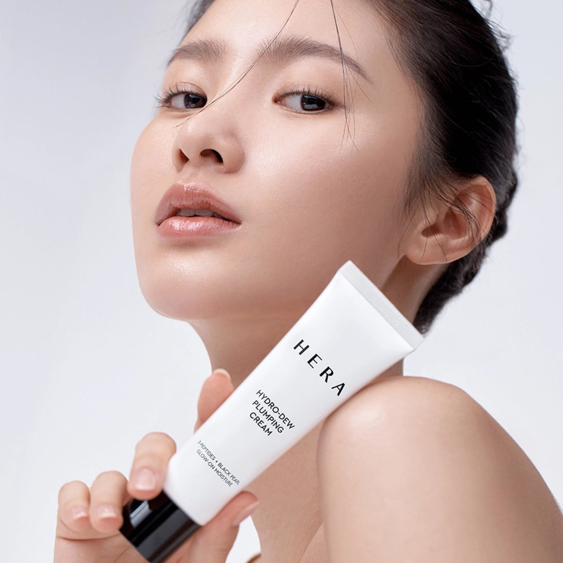 Model holding HERA Hydro-Dew Plumping Cream 50ml showcasing skincare product benefits and packaging.