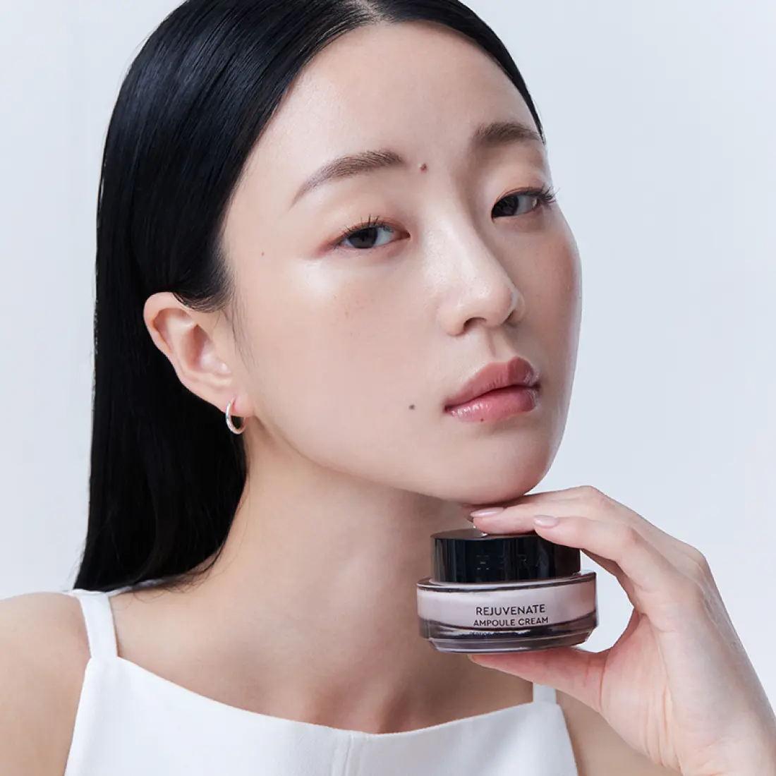 Woman holding HERA Rejuvanate Ampoule Cream 50ml jar close to her face, showcasing skincare product.