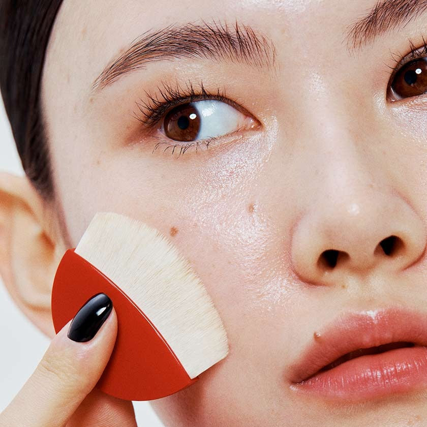Close-up of woman applying HERA Skin Tint Comfy Spreader SPF50+ PA++++ 15g with a brush, showcasing smooth, radiant skin.