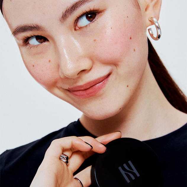 Woman holding HERA Skin Tint Comfy Spreader SPF50+ PA++++ 15g, smiling with flawless skin, highlighting product's benefits.