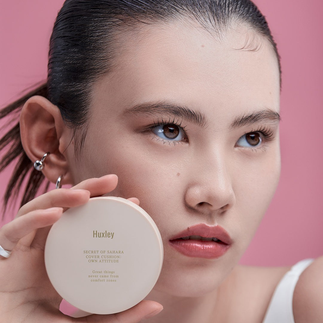 Woman holding HUXLEY Cover Cushion Own Attitude against a pink backdrop, showcasing the product's sleek design and elegance.