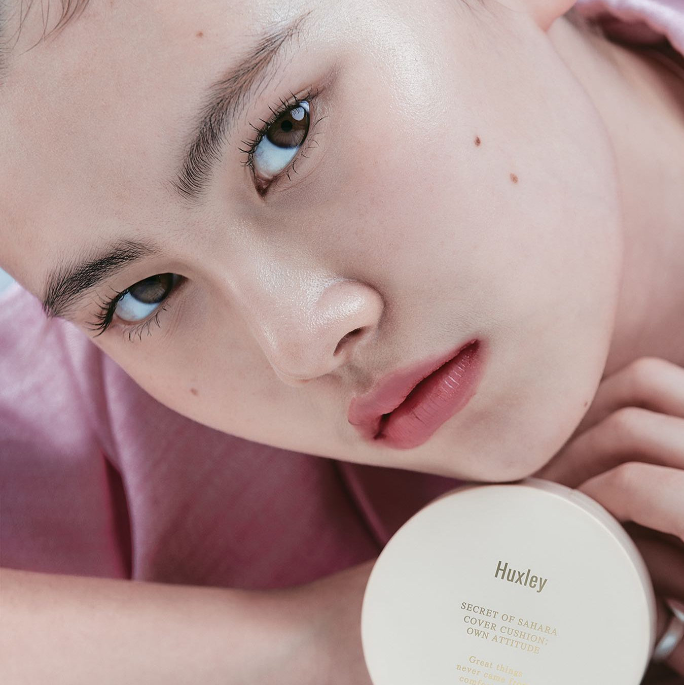 Close-up of woman holding HUXLEY Cover Cushion Own Attitude, showcasing smooth complexion and product packaging.