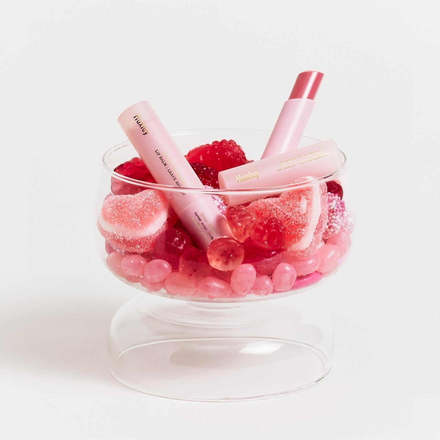HUXLEY Lip Balm Leave Behide surrounded by pink candies in a glass bowl. Enhance lip moisture and shine with HUXLEY's nourishing balm.