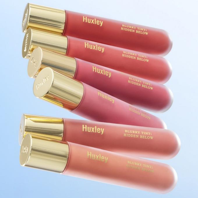 HUXLEY Blurry Tint Hidden Below lip tints in various shades displayed against a blue background with gold caps.