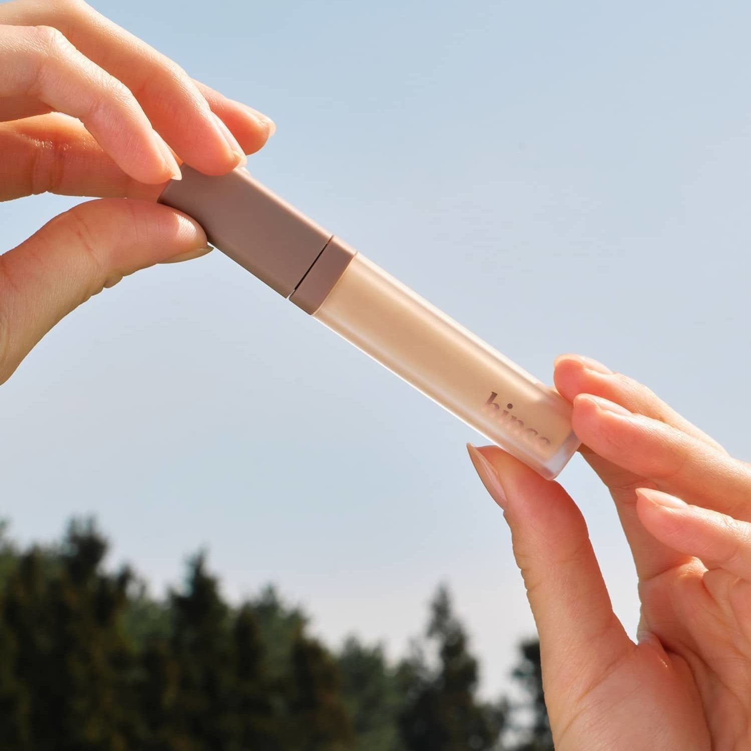 Hands holding the HINCE Second Skin Cover Concealer against a clear sky, highlighting its sleek design and natural finish.